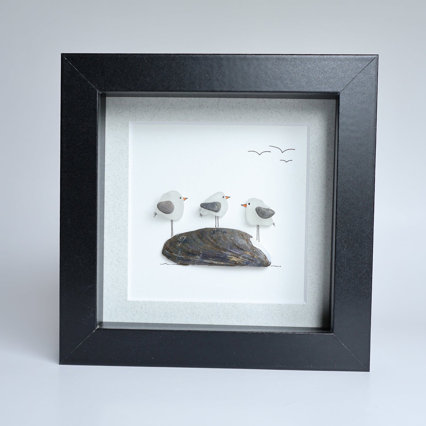 A square black shadow box frame containing a white background with three small seagull figures standing on a dark rock, with a few birds drawn in the sky.