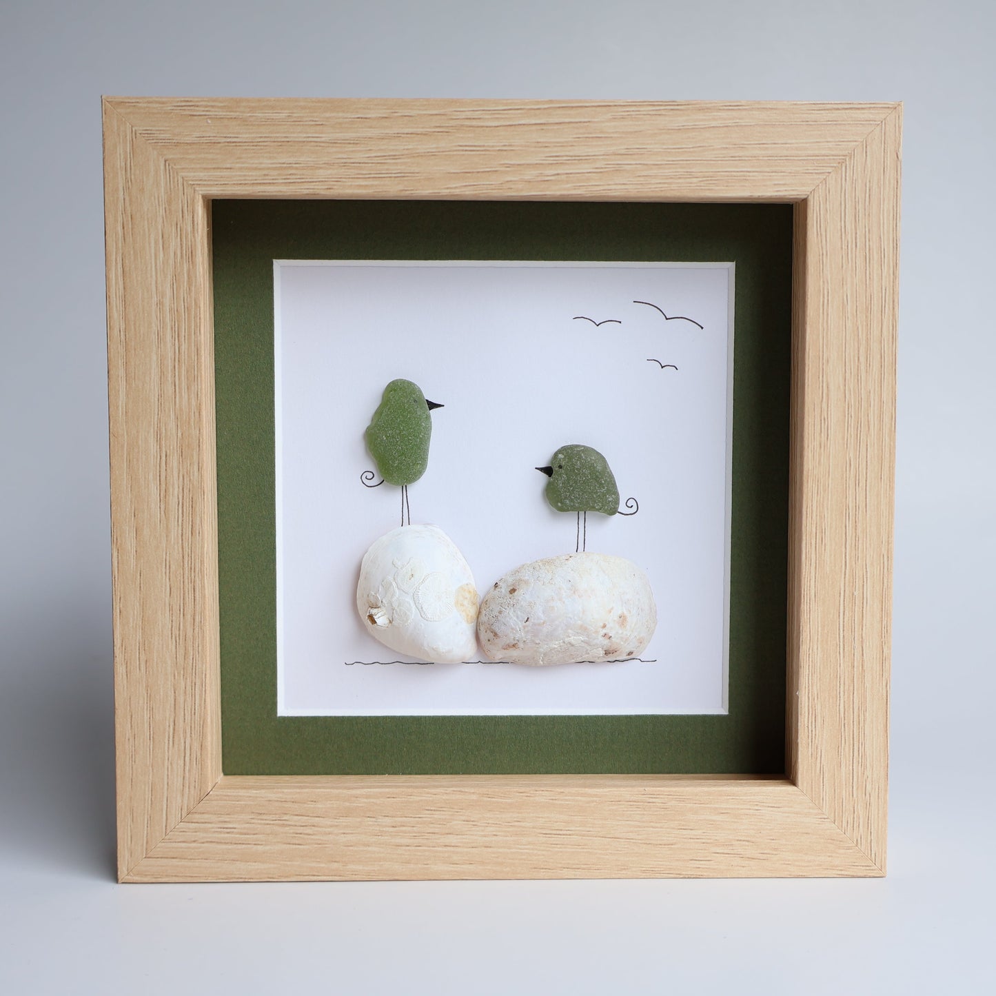 A square framed shadow box featuring two small green birds perched on white seashells, set against a white background with a dark green inner mat and a light natural wood frame.