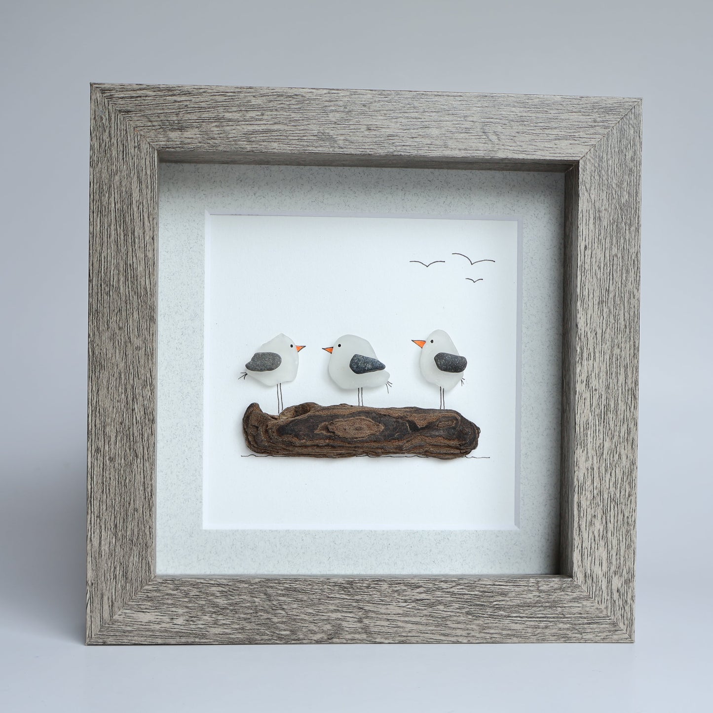 A square framed art piece featuring three small white seagulls with gray wings and orange beaks perched on a piece of driftwood, displayed against a light gray background.