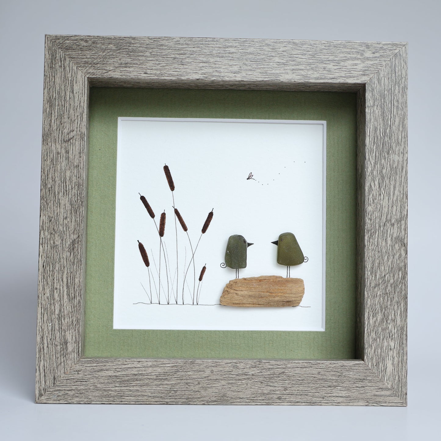 A square framed piece of handmade wall art featuring two small green bird figures on a piece of wood, with reeds on a white background and sage green matting, in a weathered gray wood frame.
