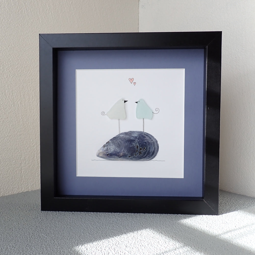 black shadowbox framed picture of two seaglass birds standing on a mussel shell