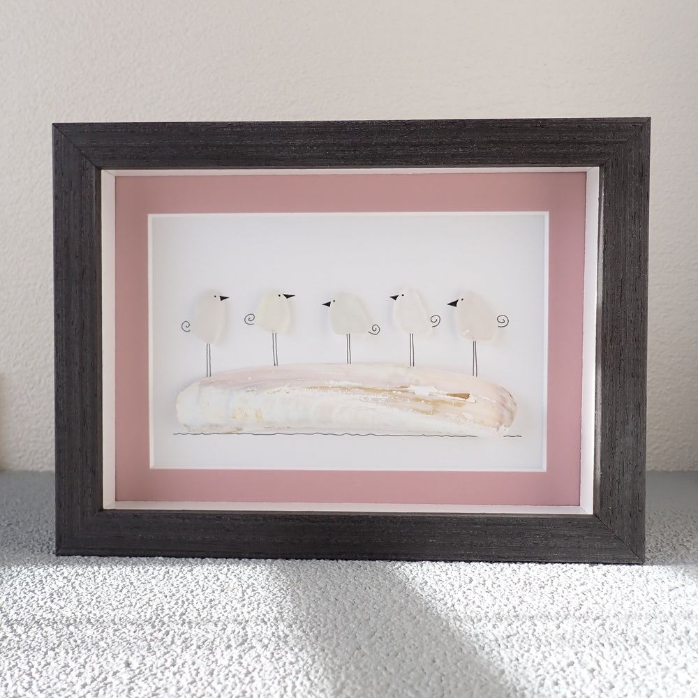 black shadowbox frame picture of a razor clam with five seaglass birds standing on it framed with a pink mat